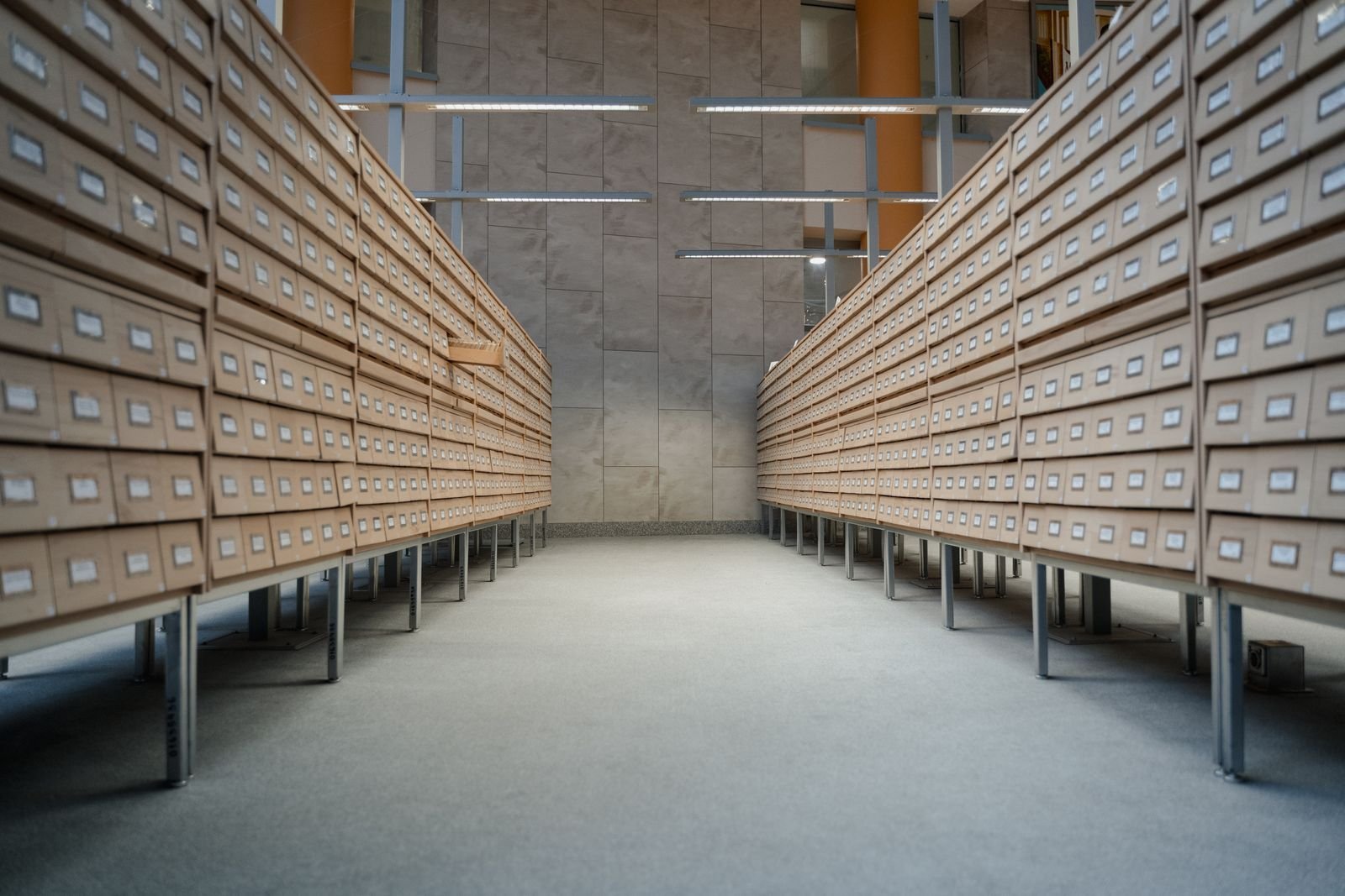 Local d'archives vide, deux longues rangées de casiers en bois clair à petits tiroirs se faisant face, sol lisse et éclairage linéaire au plafond.
