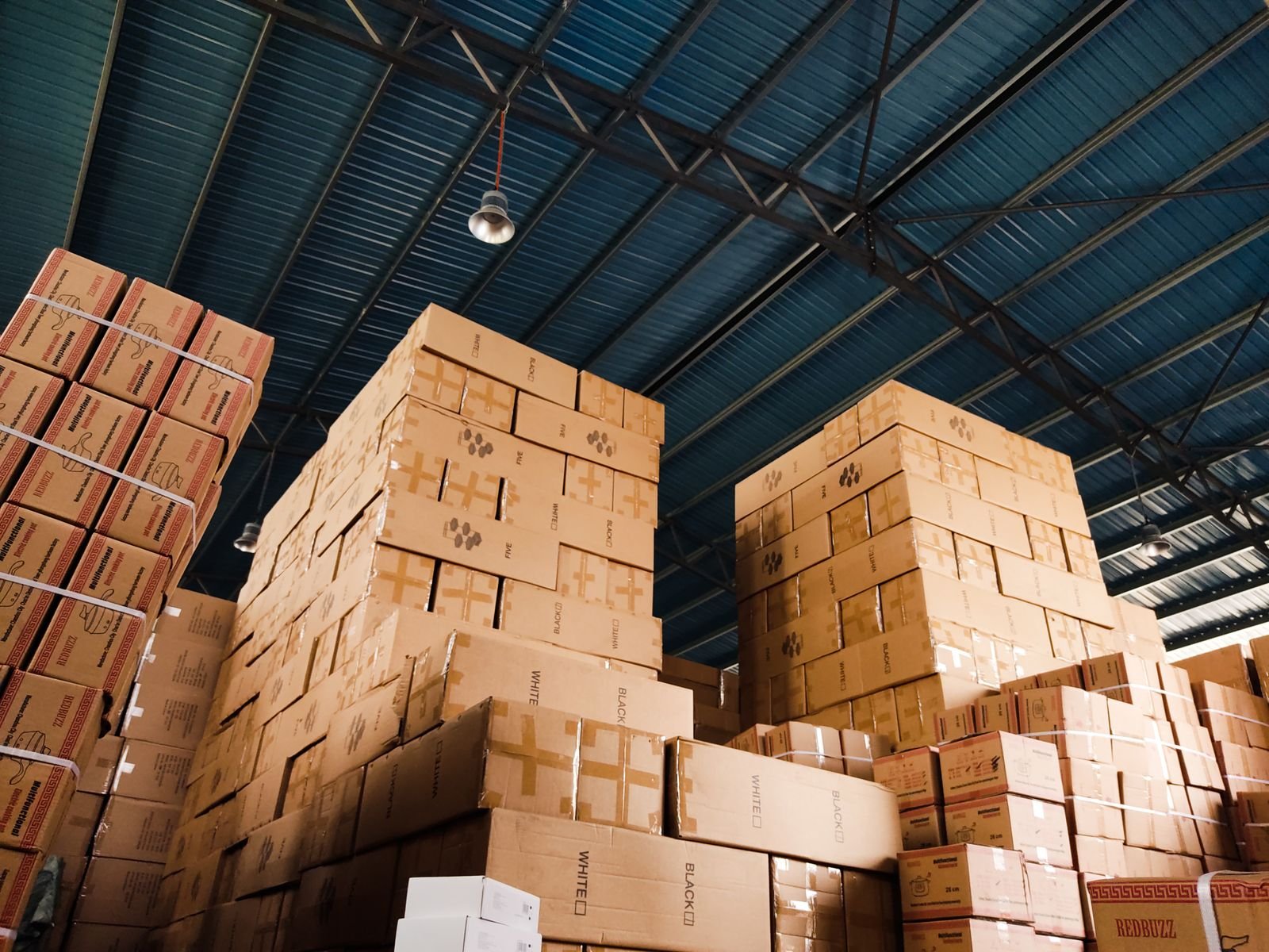 Empilements de cartons de stockage montant jusque sous une charpente métallique, vus en contre-plongée dans un local de dépôt.