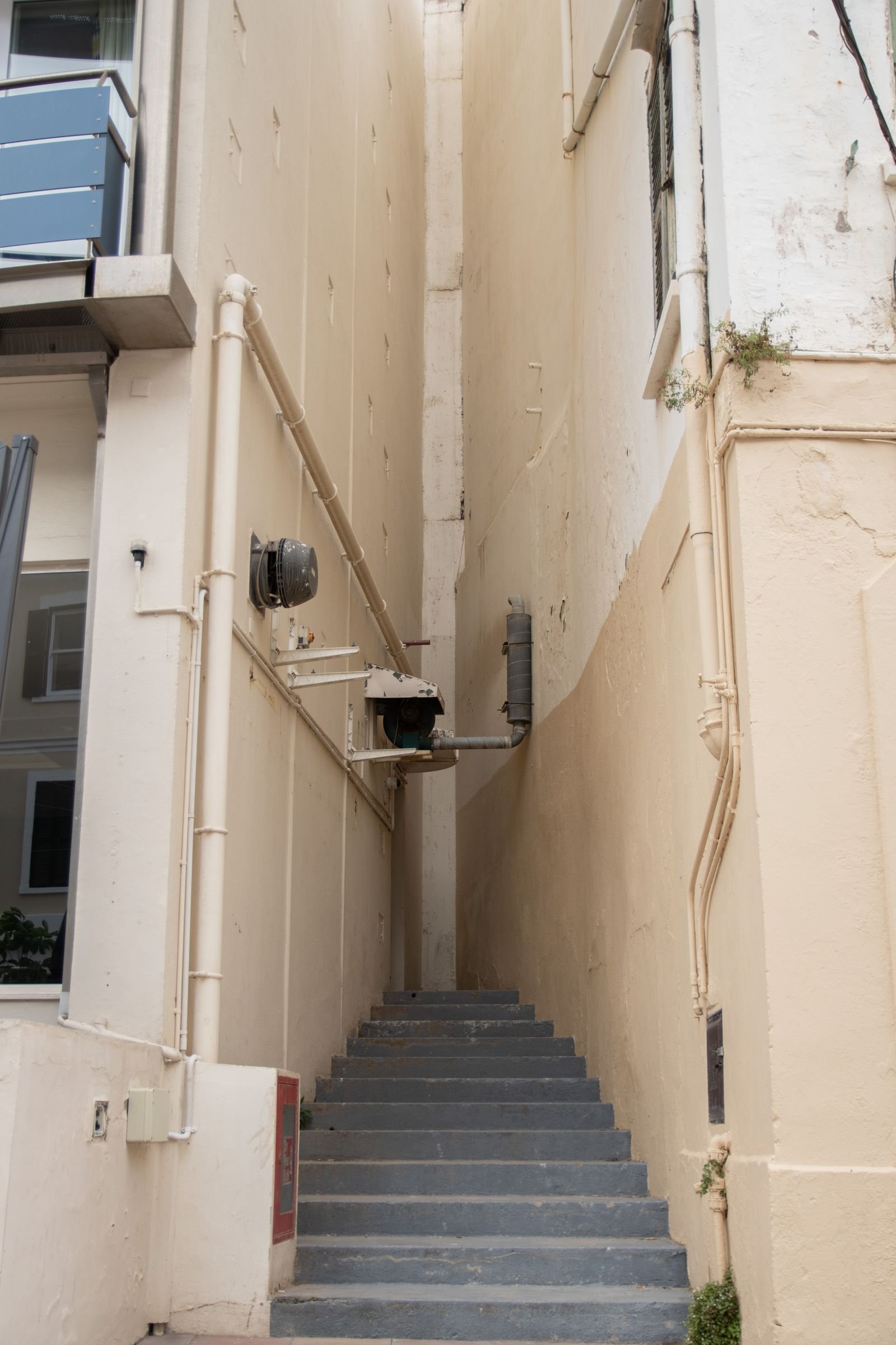 Escalier de service très étroit coincé entre deux murs clairs, marches bétonnées, coffrets techniques fixés en paroi.