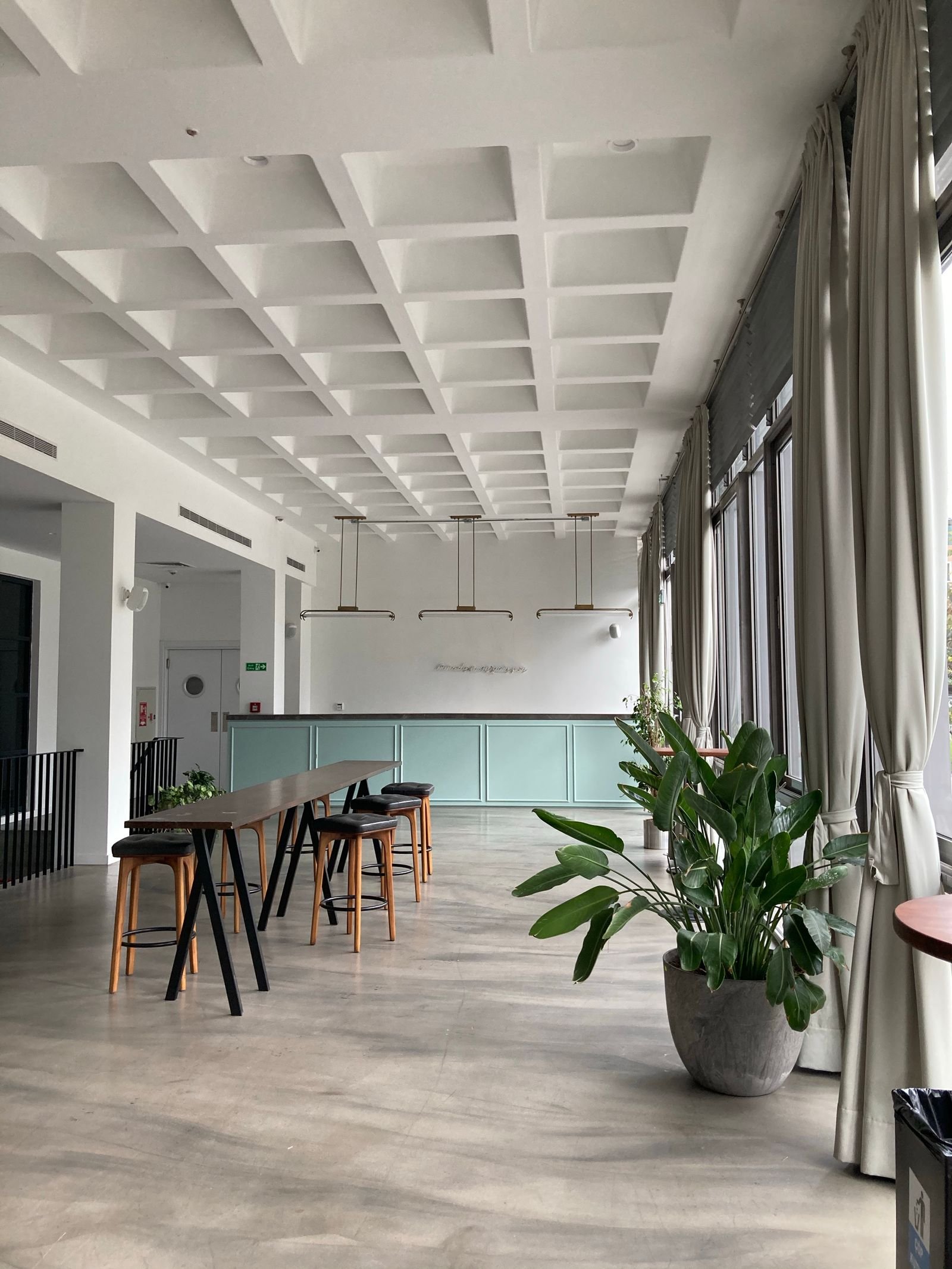 Espace de travail contemporain vide, plafond à caissons blancs, longue banque en bois, sol béton ciré et large baie vitrée voilée.