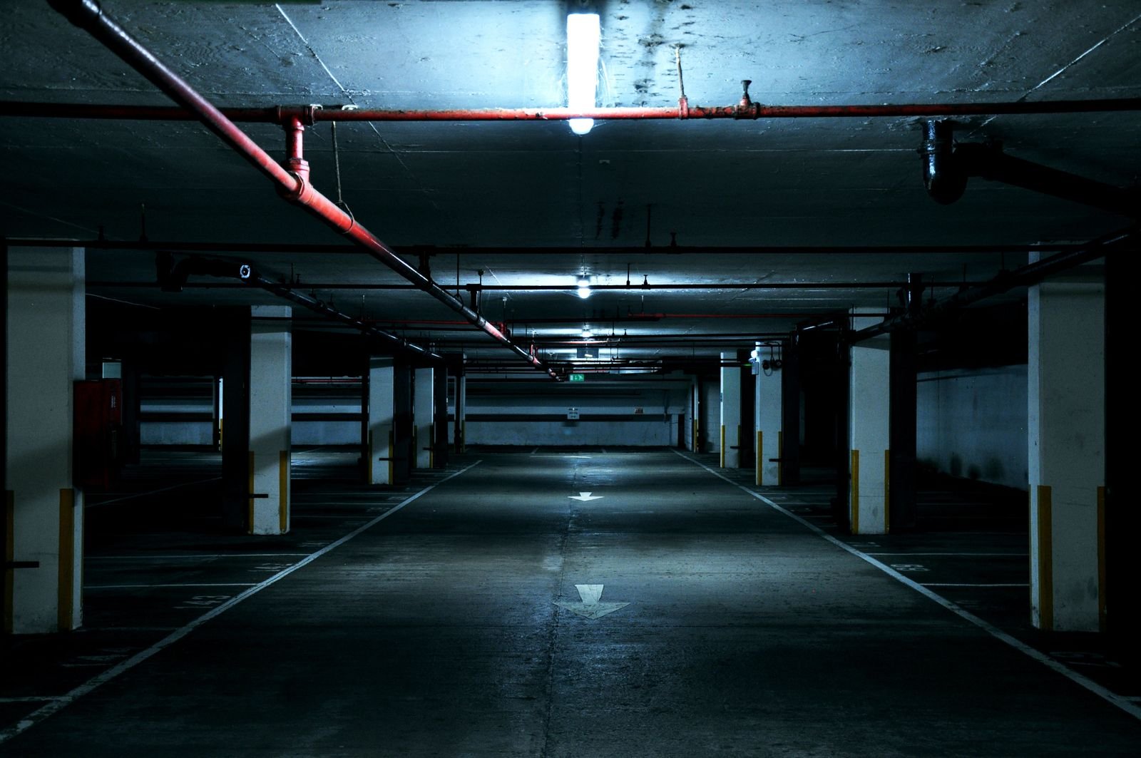 Allée centrale d'un parking souterrain désert, éclairage froid au plafond, réseaux et canalisations apparents, perspective fuyante.