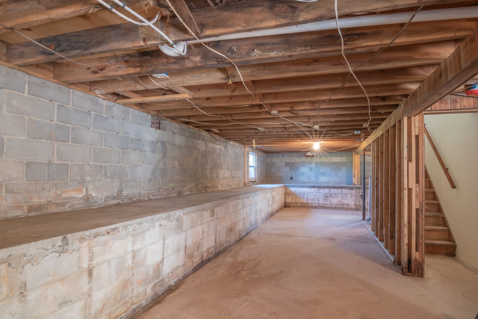 Sous-sol vide et bas de plafond, murs de parpaing et de béton portant des traces d'humidité, solives de bois apparentes et escalier au fond.